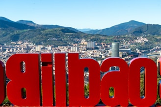 A large red sign displaying the name of a city is prominently featured with an urban cityscape in the background, set against a backdrop of rolling green hills and mountains. The city skyline includes various modern buildings and a river winding through the area.
