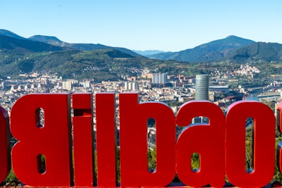A large red sign displaying the name of a city is prominently featured with an urban cityscape in the background, set against a backdrop of rolling green hills and mountains. The city skyline includes various modern buildings and a river winding through the area.