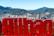 A large red sign displaying the name of a city is prominently featured with an urban cityscape in the background, set against a backdrop of rolling green hills and mountains. The city skyline includes various modern buildings and a river winding through the area.