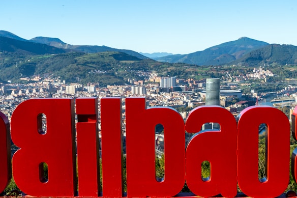 A large red sign displaying the name of a city is prominently featured with an urban cityscape in the background, set against a backdrop of rolling green hills and mountains. The city skyline includes various modern buildings and a river winding through the area.