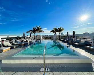 An outdoor infinity pool on a rooftop with a clear blue sky above. Palm trees are lined along the edge of the pool, and several lounge chairs are arranged on either side. The sun is shining brightly, creating a serene and inviting atmosphere.