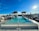 An outdoor infinity pool on a rooftop with a clear blue sky above. Palm trees are lined along the edge of the pool, and several lounge chairs are arranged on either side. The sun is shining brightly, creating a serene and inviting atmosphere.