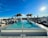 An outdoor infinity pool on a rooftop with a clear blue sky above. Palm trees are lined along the edge of the pool, and several lounge chairs are arranged on either side. The sun is shining brightly, creating a serene and inviting atmosphere.