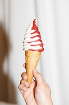A hand is holding a soft serve ice cream cone swirled with red and white colors. The ice cream has a smooth texture, and the cone appears to be a waffle type. The background is a neutral, light color.