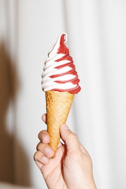 A hand is holding a soft serve ice cream cone swirled with red and white colors. The ice cream has a smooth texture, and the cone appears to be a waffle type. The background is a neutral, light color.