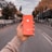 A person holds up a red iPhone with the Apple logo visible, standing in the middle of a deserted street lined with trees that have orange-brown leaves. The street is flanked by a mix of buildings and a fenced sidewalk. The sky above is overcast.
