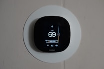 A smart thermostat is mounted on a wall, displaying the temperature set to 69 degrees. The device has a modern, sleek design with a black faceplate surrounded by a white circular backplate. Various sensor icons and settings are visible on its digital display.