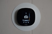 A smart thermostat is mounted on a wall, displaying the temperature set to 69 degrees. The device has a modern, sleek design with a black faceplate surrounded by a white circular backplate. Various sensor icons and settings are visible on its digital display.