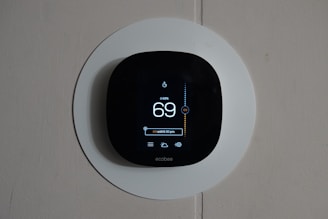 A smart thermostat is mounted on a wall, displaying the temperature set to 69 degrees. The device has a modern, sleek design with a black faceplate surrounded by a white circular backplate. Various sensor icons and settings are visible on its digital display.