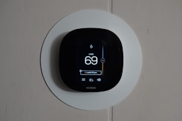 A smart thermostat is mounted on a wall, displaying the temperature set to 69 degrees. The device has a modern, sleek design with a black faceplate surrounded by a white circular backplate. Various sensor icons and settings are visible on its digital display.