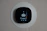 A smart thermostat is mounted on a wall, displaying the temperature set to 69 degrees. The device has a modern, sleek design with a black faceplate surrounded by a white circular backplate. Various sensor icons and settings are visible on its digital display.