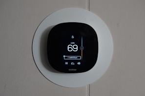 A smart thermostat is mounted on a wall, displaying the temperature set to 69 degrees. The device has a modern, sleek design with a black faceplate surrounded by a white circular backplate. Various sensor icons and settings are visible on its digital display.