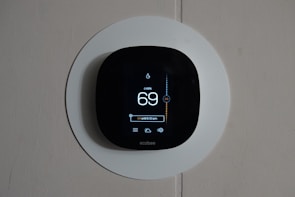 A smart thermostat is mounted on a wall, displaying the temperature set to 69 degrees. The device has a modern, sleek design with a black faceplate surrounded by a white circular backplate. Various sensor icons and settings are visible on its digital display.