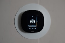 A smart thermostat is mounted on a wall, displaying the temperature set to 69 degrees. The device has a modern, sleek design with a black faceplate surrounded by a white circular backplate. Various sensor icons and settings are visible on its digital display.