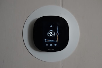 A smart thermostat is mounted on a wall, displaying the temperature set to 69 degrees. The device has a modern, sleek design with a black faceplate surrounded by a white circular backplate. Various sensor icons and settings are visible on its digital display.