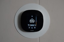 A smart thermostat is mounted on a wall, displaying the temperature set to 69 degrees. The device has a modern, sleek design with a black faceplate surrounded by a white circular backplate. Various sensor icons and settings are visible on its digital display.