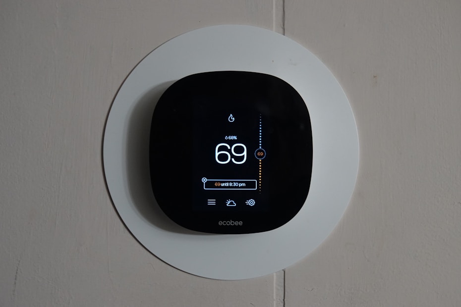 A smart thermostat is mounted on a wall, displaying the temperature set to 69 degrees. The device has a modern, sleek design with a black faceplate surrounded by a white circular backplate. Various sensor icons and settings are visible on its digital display.