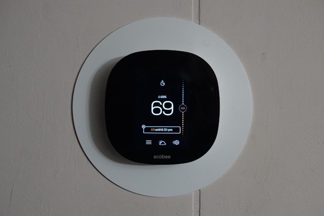 A smart thermostat is mounted on a wall, displaying the temperature set to 69 degrees. The device has a modern, sleek design with a black faceplate surrounded by a white circular backplate. Various sensor icons and settings are visible on its digital display.