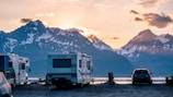 A scenic landscape with several recreational vehicles (RVs) parked near a calm body of water. Snow-capped mountains are visible in the background, and the sky is painted with the warm hues of either sunrise or sunset. A car is also parked near the RVs, and there is a sense of tranquility and natural beauty.