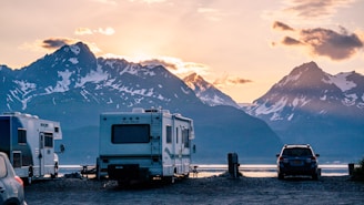 A scenic landscape with several recreational vehicles (RVs) parked near a calm body of water. Snow-capped mountains are visible in the background, and the sky is painted with the warm hues of either sunrise or sunset. A car is also parked near the RVs, and there is a sense of tranquility and natural beauty.