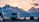 A scenic landscape with several recreational vehicles (RVs) parked near a calm body of water. Snow-capped mountains are visible in the background, and the sky is painted with the warm hues of either sunrise or sunset. A car is also parked near the RVs, and there is a sense of tranquility and natural beauty.