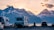 A scenic landscape with several recreational vehicles (RVs) parked near a calm body of water. Snow-capped mountains are visible in the background, and the sky is painted with the warm hues of either sunrise or sunset. A car is also parked near the RVs, and there is a sense of tranquility and natural beauty.