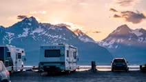 A scenic landscape with several recreational vehicles (RVs) parked near a calm body of water. Snow-capped mountains are visible in the background, and the sky is painted with the warm hues of either sunrise or sunset. A car is also parked near the RVs, and there is a sense of tranquility and natural beauty.