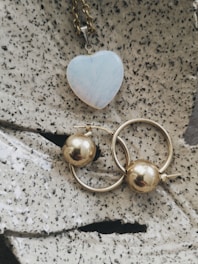 A heart-shaped pendant with a pale iridescent surface is attached to a gold chain, placed on a textured stone-like surface. Below it, two gold hoop earrings with spherical gold beads are positioned prominently.