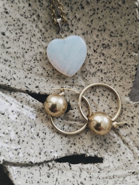 A heart-shaped pendant with a pale iridescent surface is attached to a gold chain, placed on a textured stone-like surface. Below it, two gold hoop earrings with spherical gold beads are positioned prominently.