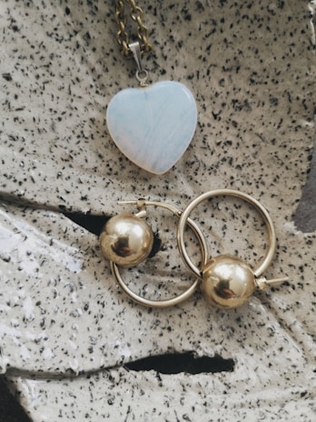 A heart-shaped pendant with a pale iridescent surface is attached to a gold chain, placed on a textured stone-like surface. Below it, two gold hoop earrings with spherical gold beads are positioned prominently.