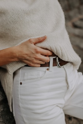 A person wearing a cozy cream sweater paired with white jeans, holding a brown leather belt with their right hand.