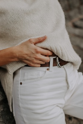A person wearing a cozy cream sweater paired with white jeans, holding a brown leather belt with their right hand.