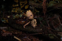 A small, tree-like creature with large eyes and a gentle expression sits on a mossy log in a dense forest. Surrounding the creature are fallen leaves and various shades of green foliage, creating a natural and vibrant setting.