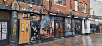 A row of buildings features a club or bar named The Bootlegger, showcasing murals and signs advertising cocktails and live music. The mural to the right includes artwork of musicians and vibrant imagery. The establishment exhibits a vintage style with dark brick and wood elements, and the pavement is wet, indicating recent rain.