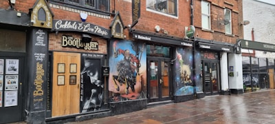 A row of buildings features a club or bar named The Bootlegger, showcasing murals and signs advertising cocktails and live music. The mural to the right includes artwork of musicians and vibrant imagery. The establishment exhibits a vintage style with dark brick and wood elements, and the pavement is wet, indicating recent rain.