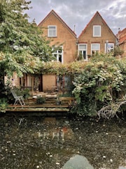 Two quaint brick houses with gabled roofs are situated by a canal. The façade is partially covered with green foliage and vines, creating a natural and serene atmosphere. A small wooden deck with a table, chairs, and potted plants is present in front of the houses, overlooking the calm water of the canal.