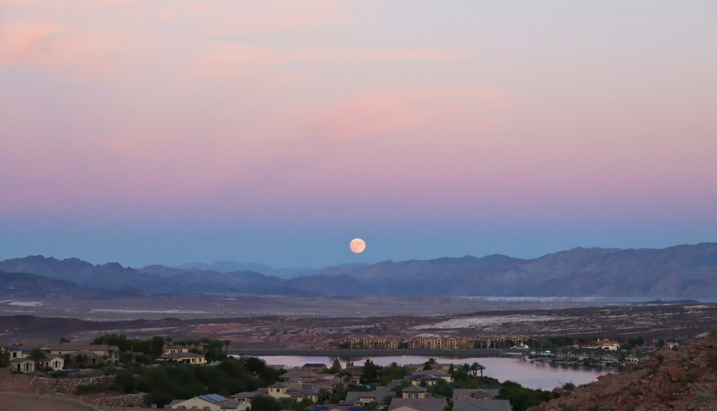 A full moon rising over a desert town ringed by mountains at dusk