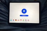 A tablet displaying a VPN application interface with a white background. The screen shows a large blue button labeled 'Disconnect' in the center, surrounded by circular lines. Below, there are several country flag icons representing different server locations, including Netherlands, Germany, Canada, Singapore, India, New York, and San Francisco. The tablet is resting on a person's lap, wearing blue jeans.