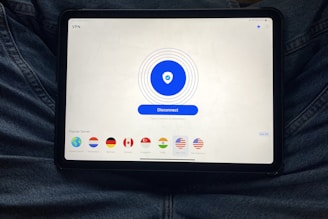 A tablet displaying a VPN application interface with a white background. The screen shows a large blue button labeled 'Disconnect' in the center, surrounded by circular lines. Below, there are several country flag icons representing different server locations, including Netherlands, Germany, Canada, Singapore, India, New York, and San Francisco. The tablet is resting on a person's lap, wearing blue jeans.