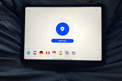 A tablet displaying a VPN application interface with a white background. The screen shows a large blue button labeled 'Disconnect' in the center, surrounded by circular lines. Below, there are several country flag icons representing different server locations, including Netherlands, Germany, Canada, Singapore, India, New York, and San Francisco. The tablet is resting on a person's lap, wearing blue jeans.