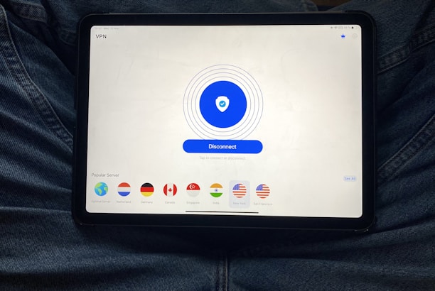A tablet displaying a VPN application interface with a white background. The screen shows a large blue button labeled 'Disconnect' in the center, surrounded by circular lines. Below, there are several country flag icons representing different server locations, including Netherlands, Germany, Canada, Singapore, India, New York, and San Francisco. The tablet is resting on a person's lap, wearing blue jeans.