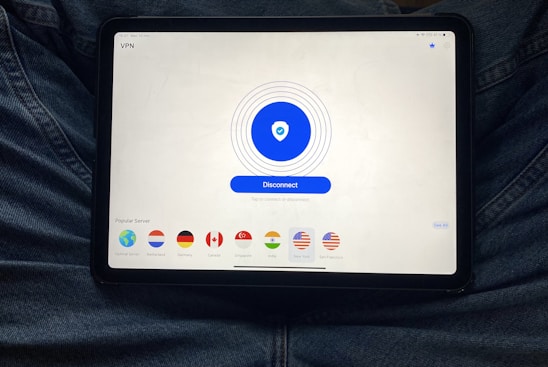A tablet displaying a VPN application interface with a white background. The screen shows a large blue button labeled 'Disconnect' in the center, surrounded by circular lines. Below, there are several country flag icons representing different server locations, including Netherlands, Germany, Canada, Singapore, India, New York, and San Francisco. The tablet is resting on a person's lap, wearing blue jeans.