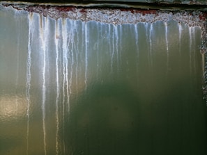 The image features a green metallic surface with noticeable streaks of rust and white corrosion lines descending vertically. The surface texture is rough, and the corroded areas display a contrast between the dark rust and the lighter spots.