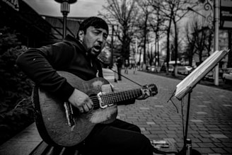 A man is sitting on a bench playing a worn-out acoustic guitar. He appears to be singing with an expressive face. There is a stand with sheet music in front of him, and the setting is an urban street lined with trees and blurred people in the background.