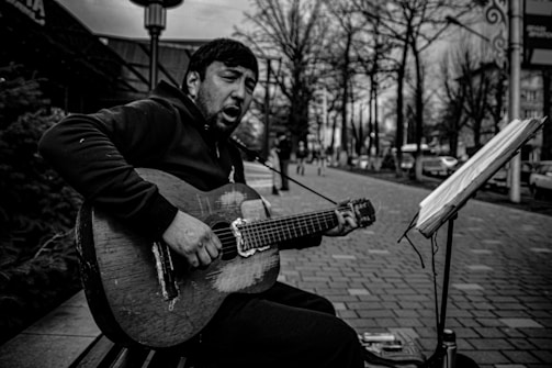 A man is sitting on a bench playing a worn-out acoustic guitar. He appears to be singing with an expressive face. There is a stand with sheet music in front of him, and the setting is an urban street lined with trees and blurred people in the background.