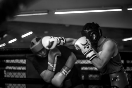 Two individuals are engaged in a boxing match inside a ring. Both are wearing protective headgear and gloves. One boxer is delivering a punch while the other attempts to block or avoid the hit. The scene is captured in black and white, adding a dramatic effect.