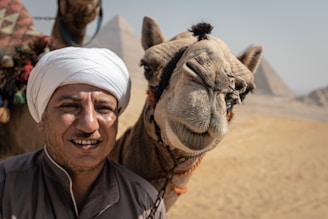 A smiling man wearing a white turban and traditional clothing stands next to a camel with a humorous expression. In the background, pyramids rise from the desert landscape.