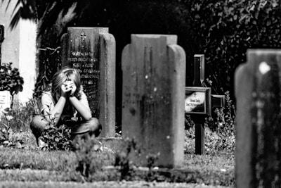 A person, appearing to be a child, is sitting in a graveyard surrounded by tombstones. The individual has their hands covering their face, suggesting distress or contemplation. The environment is somber and serene, with grass and plants growing around the gravestones.
