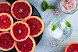 Slices of red grapefruit are arranged on a black plate, displayed beside a glass of water with ice and mint leaves. Pink rock salt is scattered on the gray surface, adding to the aesthetic appeal. A sprig of mint rests nearby the plate.