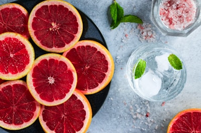 Slices of red grapefruit are arranged on a black plate, displayed beside a glass of water with ice and mint leaves. Pink rock salt is scattered on the gray surface, adding to the aesthetic appeal. A sprig of mint rests nearby the plate.